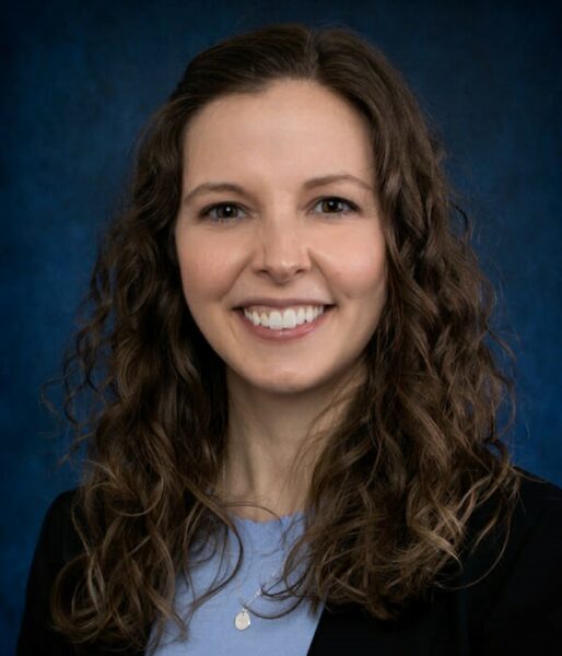 Professional headshot of a woman with curly brown hair smiling, dressed in a blue blouse against a blue background.