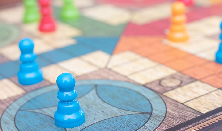 Close-up of colorful plastic game pieces on a Ludo board, showing blue, orange, green, and red playing pieces arranged on various squares.