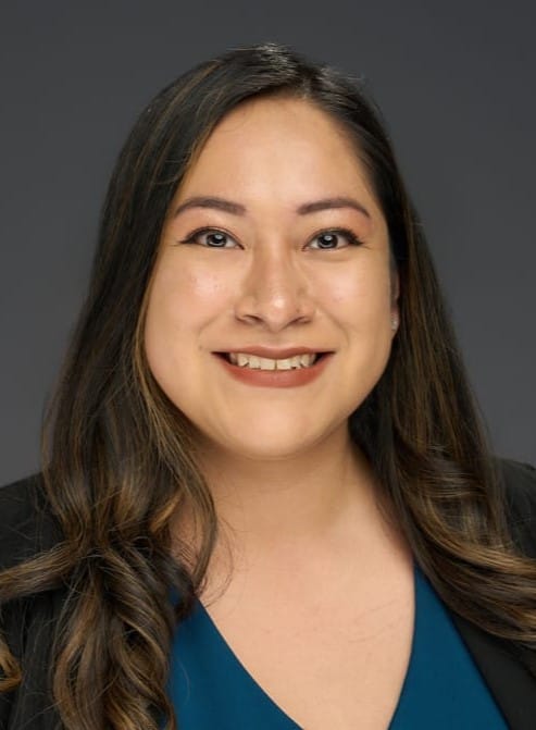 Person with long dark hair, smiling, wearing a dark blazer and a blue top against a gray background.