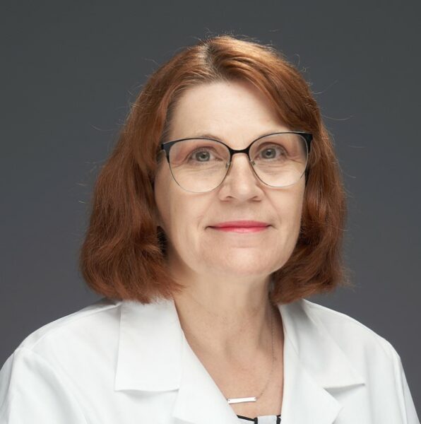 Portrait of a mature woman with shoulder-length brown hair, glasses, wearing a white lab coat, smiling at the camera against a gray background.