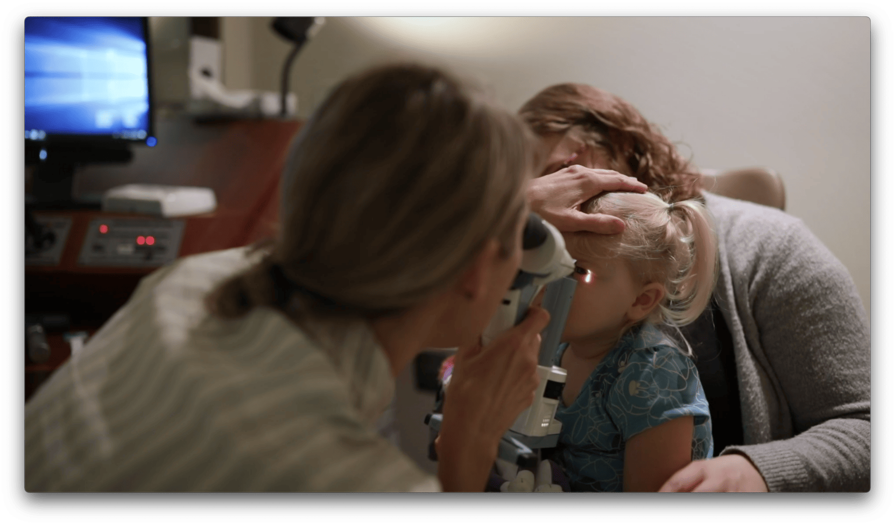 A doctor examines a young girl's eye with a medical instrument in an office setting. Another person holds the child's head steady.