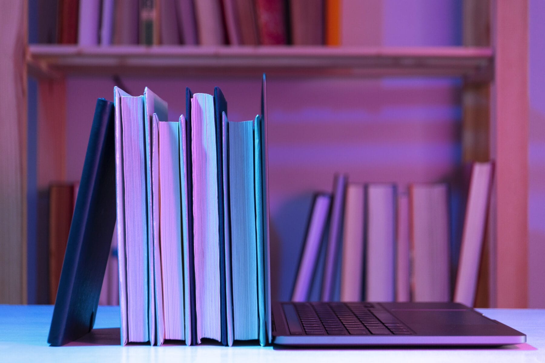 A row of books stands upright on a desk beside an open laptop, with a bookshelf filled with books in the background.