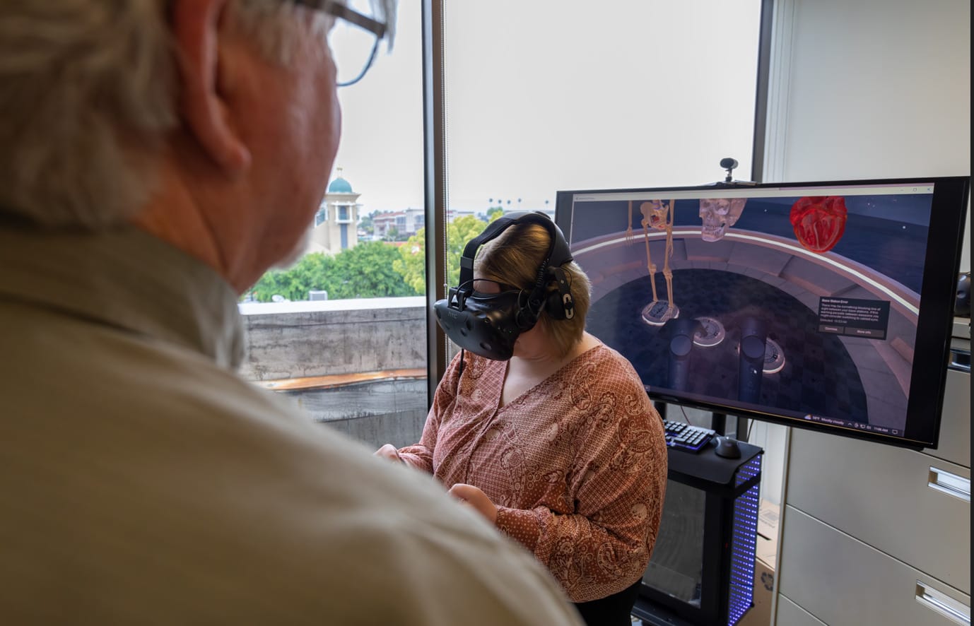 A person wearing a VR headset interacts with a virtual anatomy display on a screen while another person observes in an office setting.
