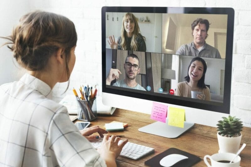 A woman sits at a desk using a computer for a video call with four people, each visible in separate windows on the monitor.