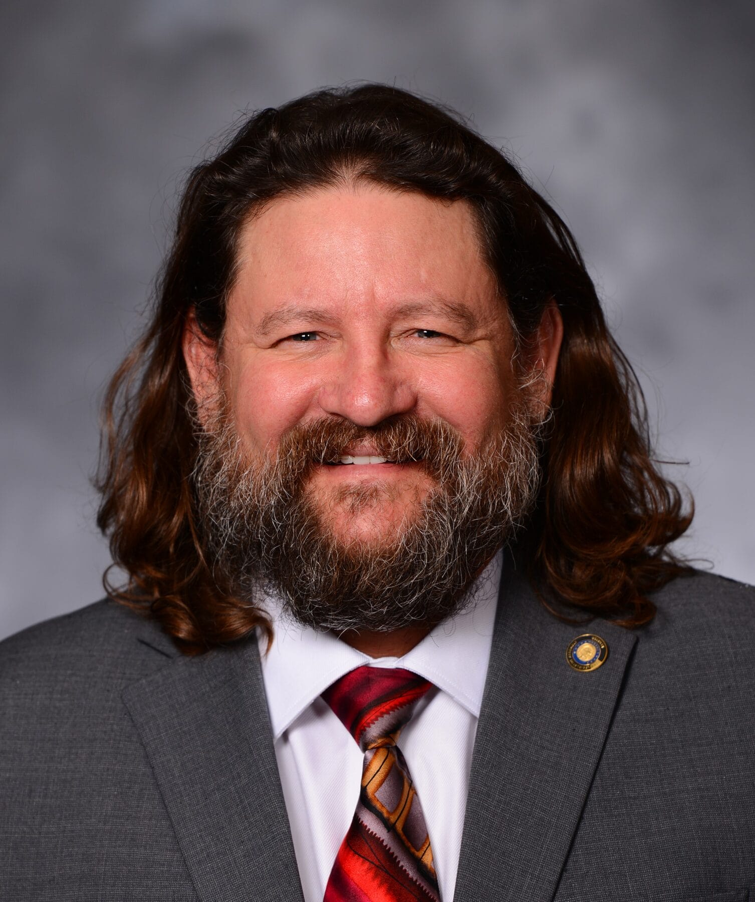 A person with long hair and a beard, wearing a gray suit, white shirt, and a red patterned tie, smiles in front of a gray background.