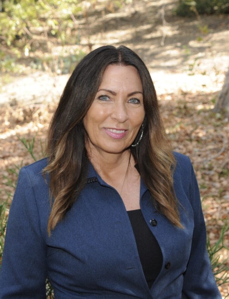 Woman with long brown hair smiling, wearing a blue jacket and standing outdoors with trees and fallen leaves in the background.