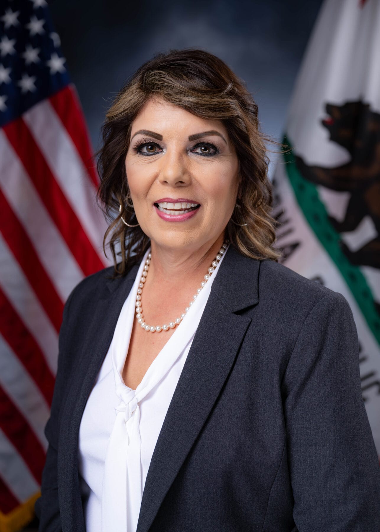 A woman in a gray blazer and white blouse stands in front of U.S. and California flags.