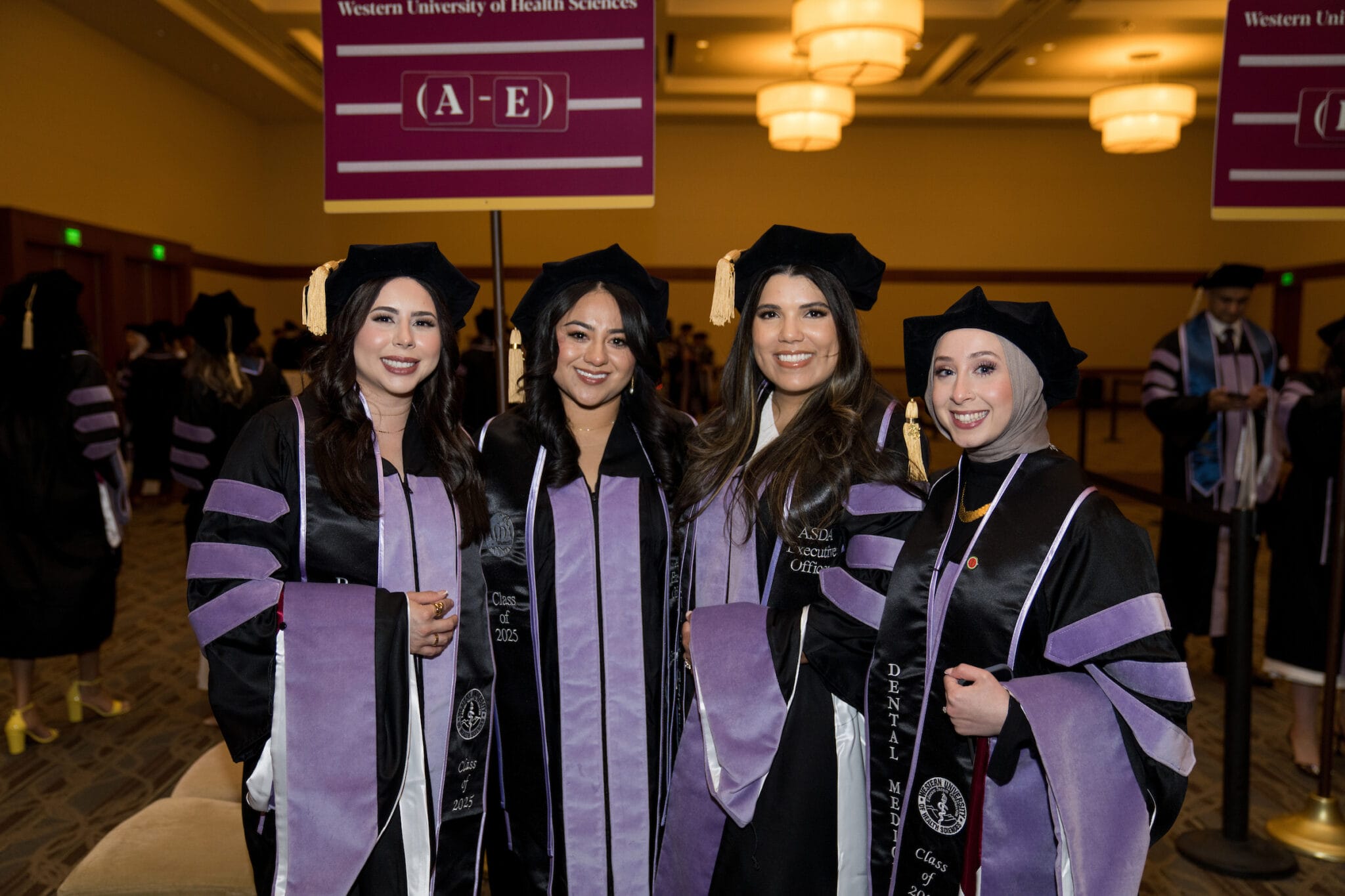 Four graduates in caps and gowns pose together at a commencement ceremony. A sign above them reads "Western University of Health Sciences A-E.