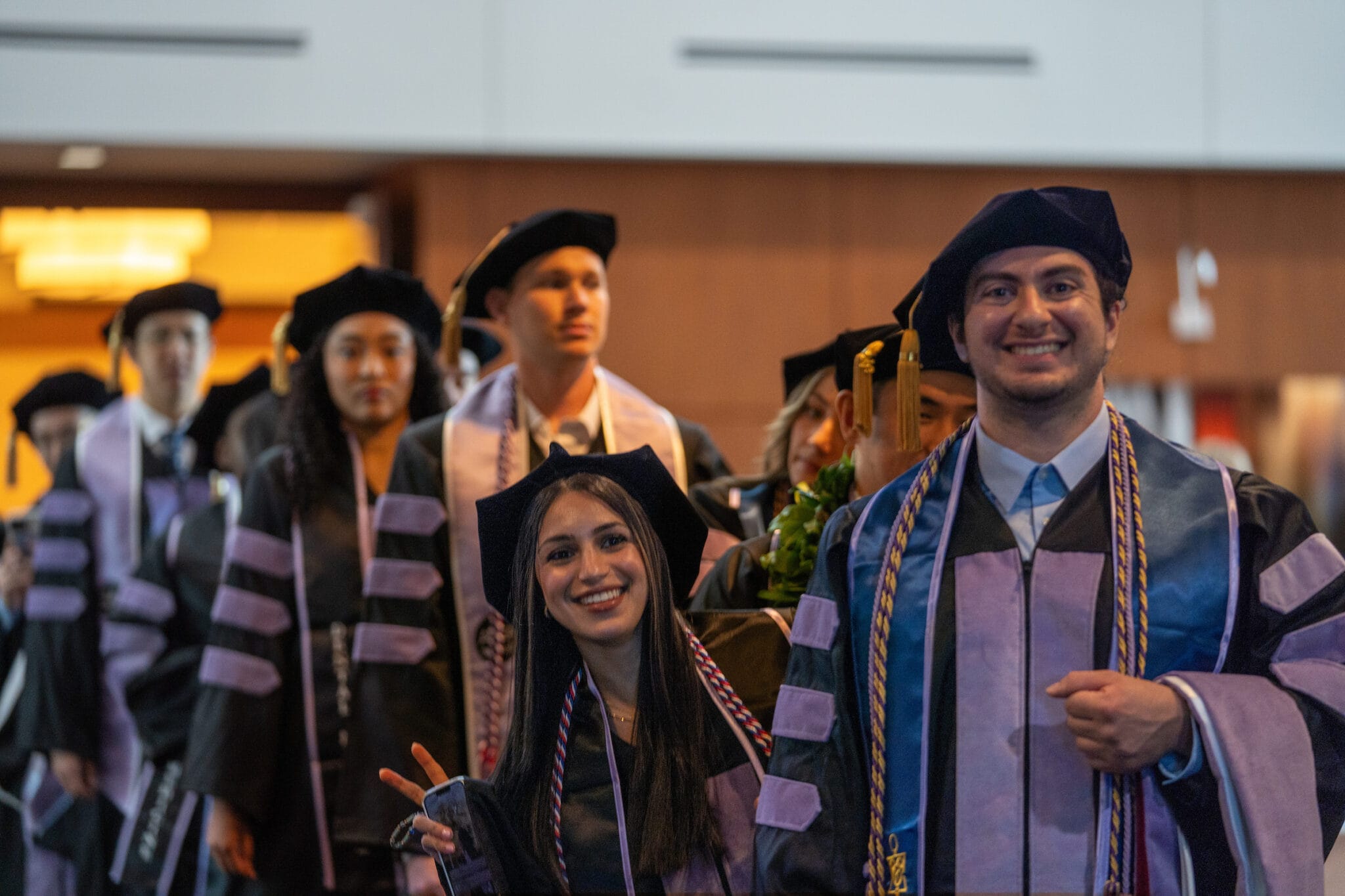 A group of graduates in caps and gowns stand in line indoors, with two smiling individuals at the front, one showing a peace sign.