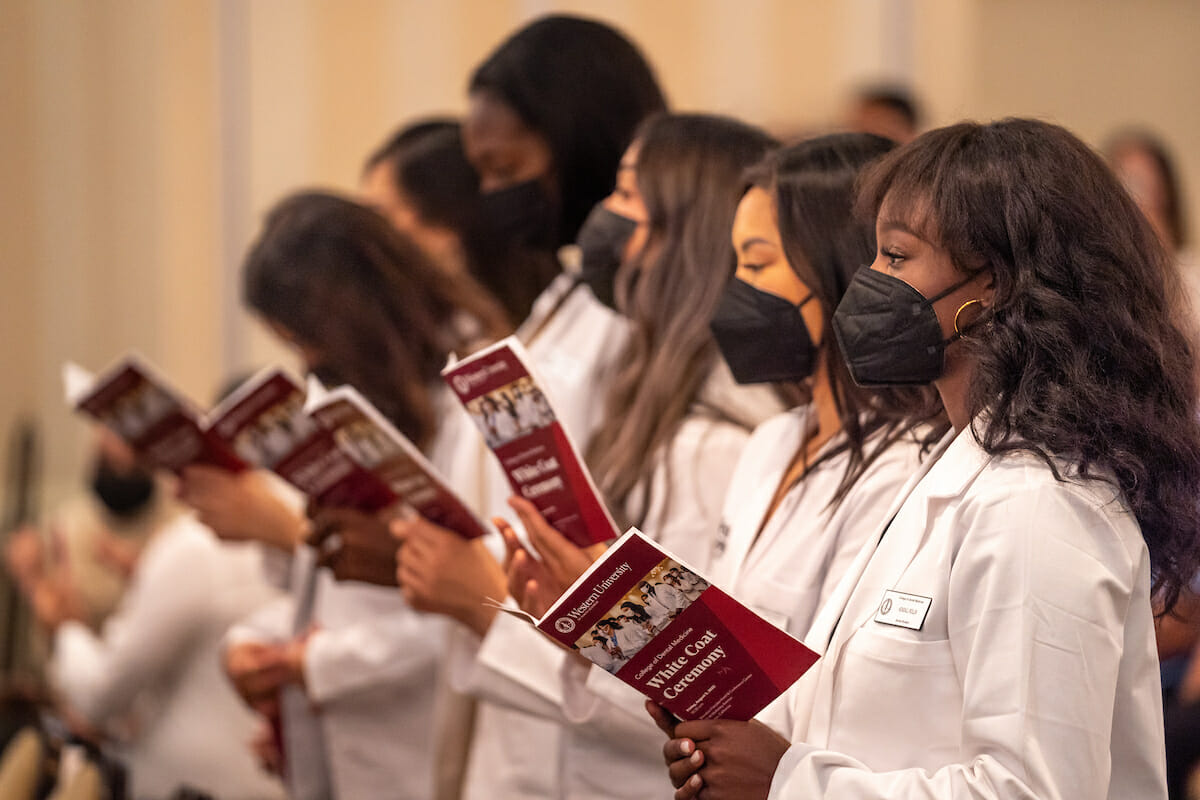 Students in white coats and wearing face masks reading from a booklet during white coat ceremony