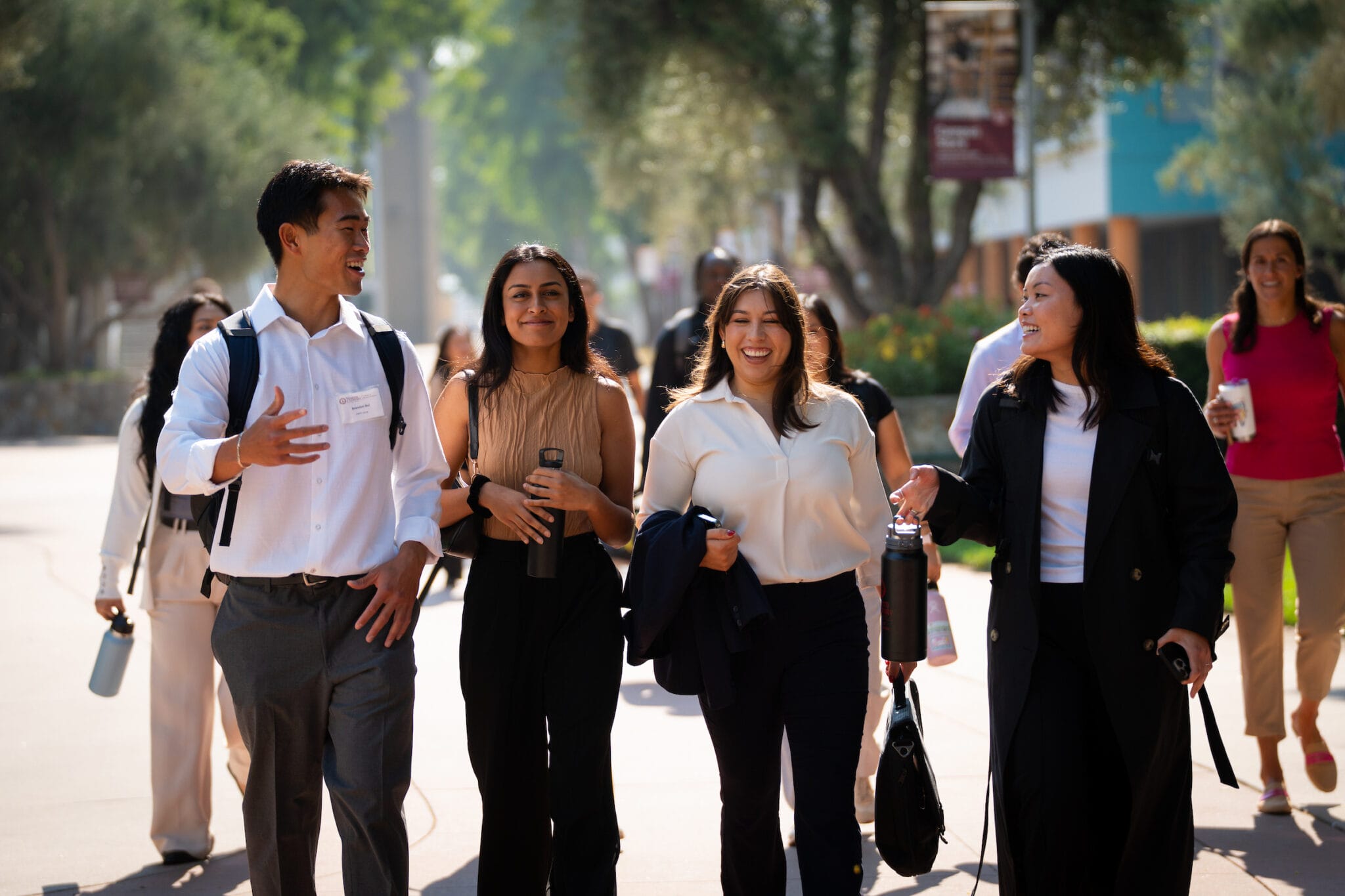 A group of professionally dressed people walk outdoors on a sunny day, talking and smiling, with trees and buildings in the background.