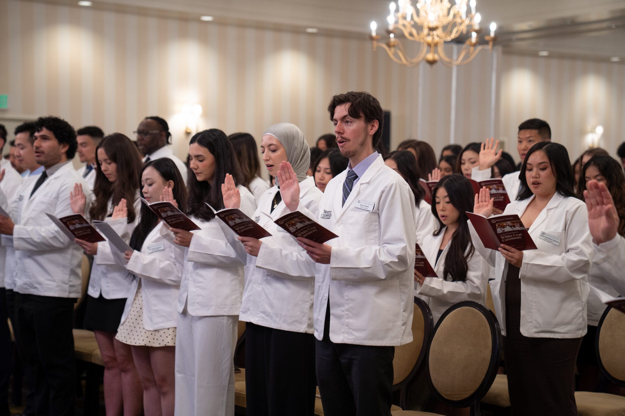 A group of people in white coats stands with raised right hands and holds booklets, participating in a formal ceremony in a well-lit room.