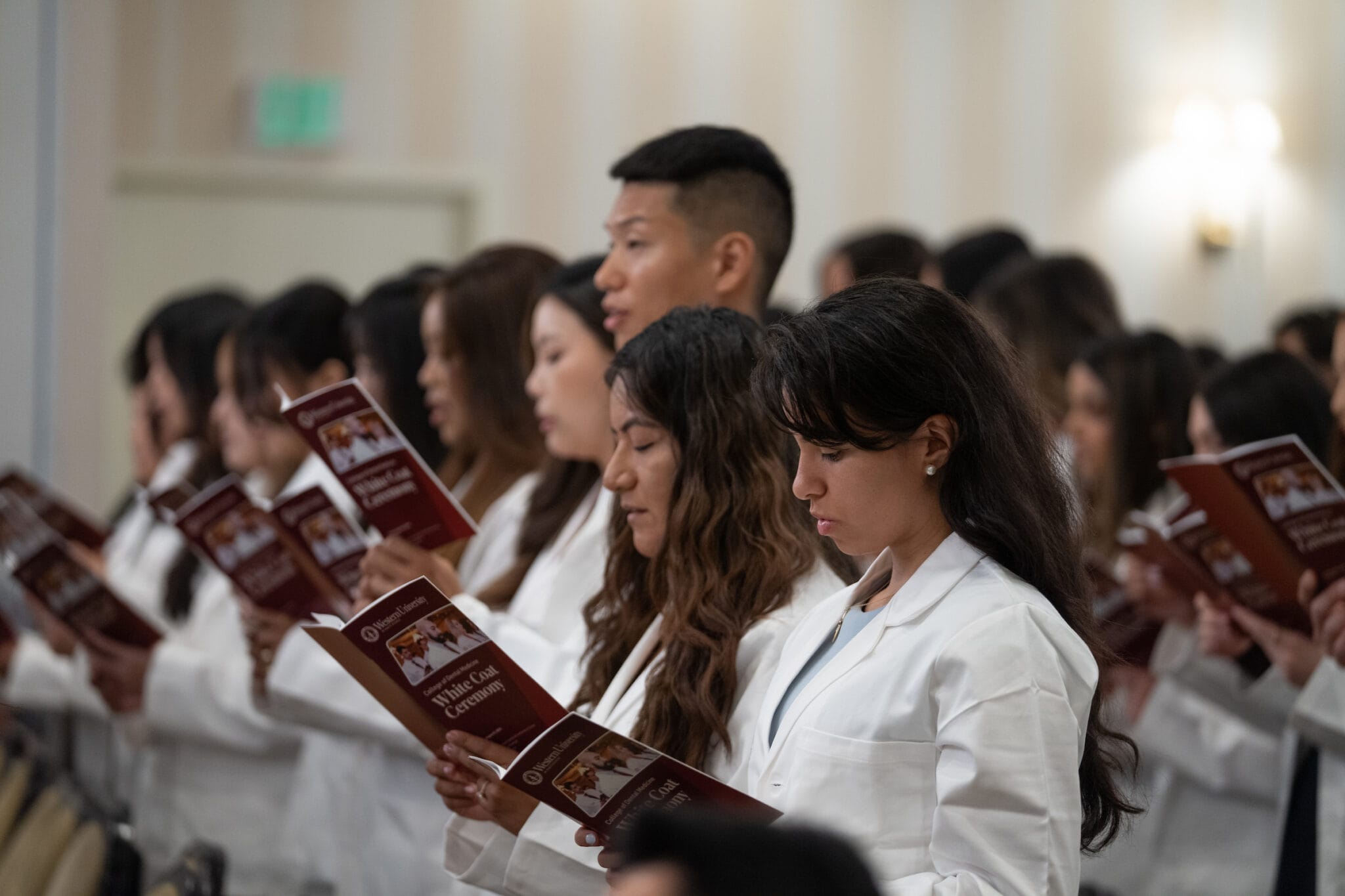 A group of people in white coats stand in rows reading from booklets during an indoor ceremony.