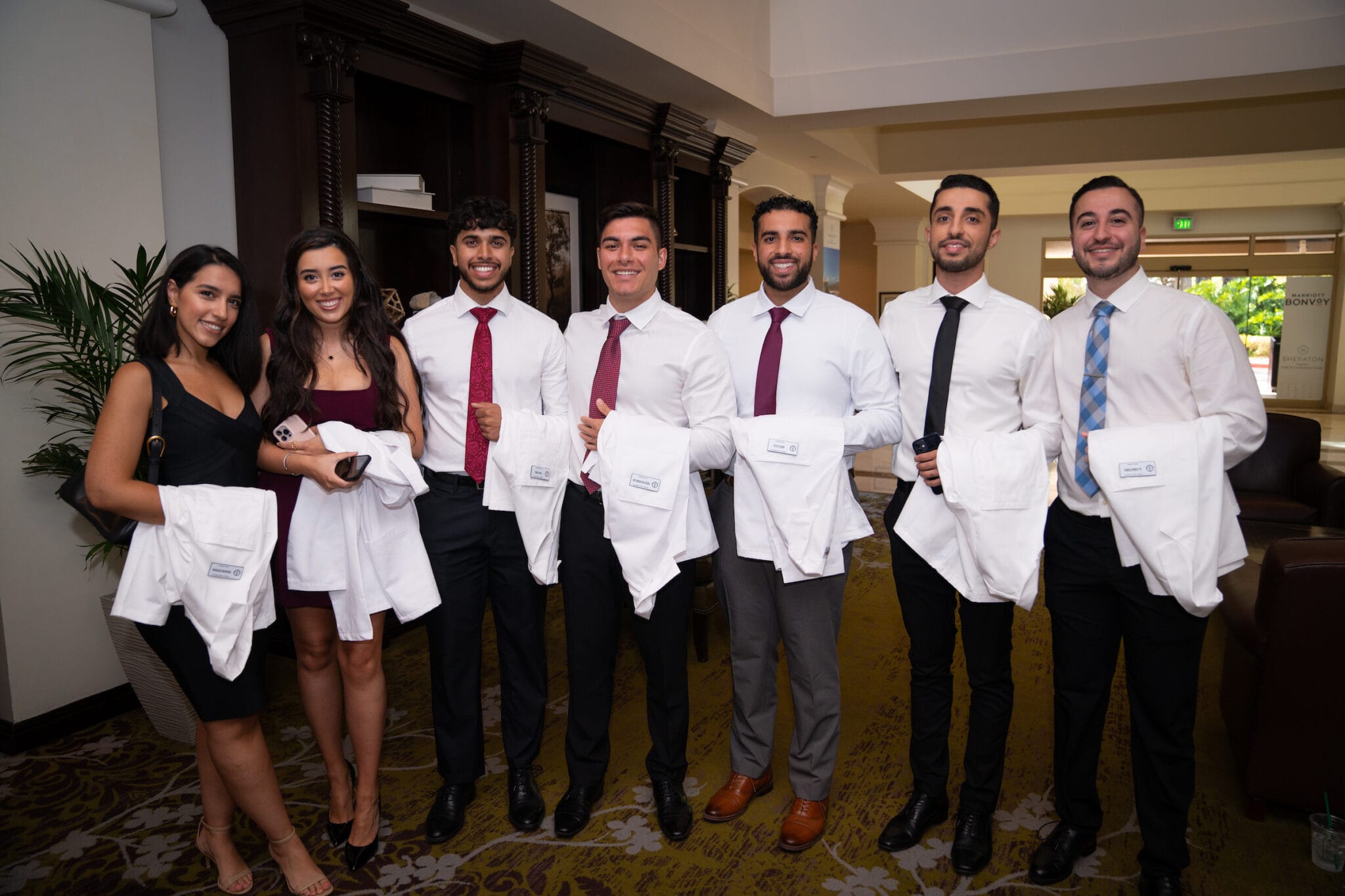 Seven young adults dressed in formal attire stand indoors, smiling at the camera; six hold white coats labeled “College of Pharmacy.”.