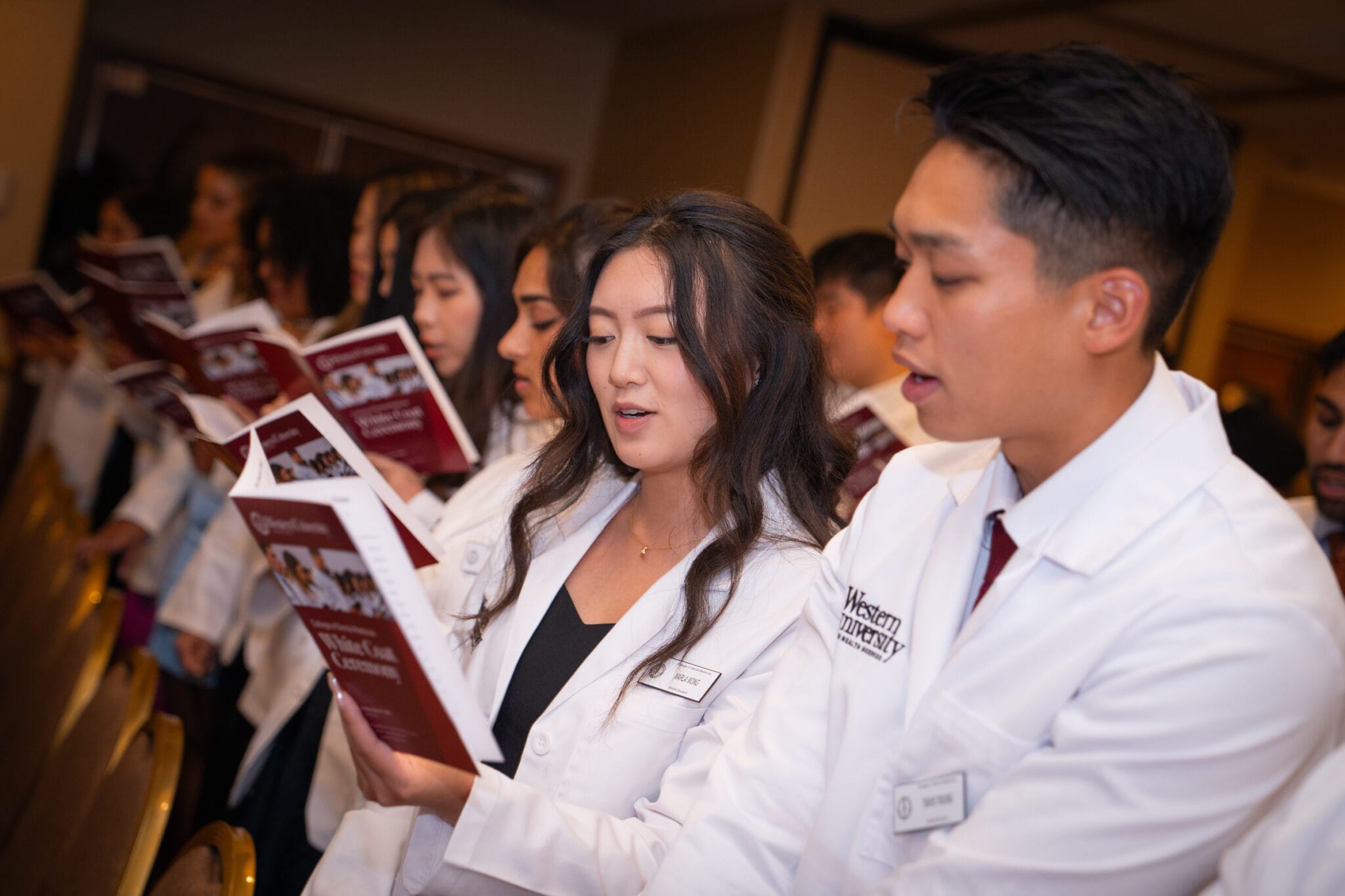 A group of students in white coats stand in a row, holding and reading from booklets at what appears to be a medical or pharmacy school ceremony.