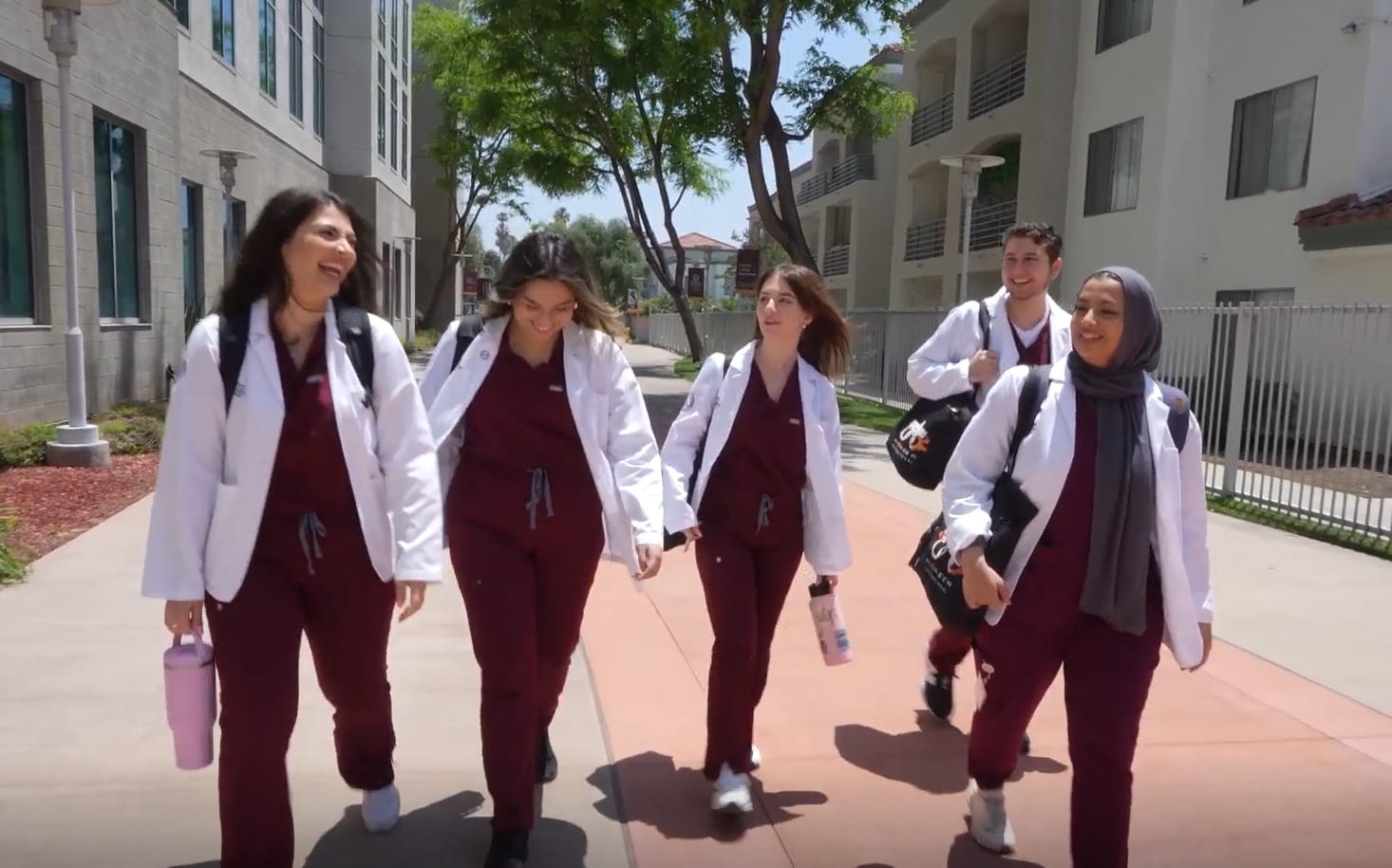 Five people in medical scrubs and white coats walking on a sidewalk between buildings, smiling and carrying bags.