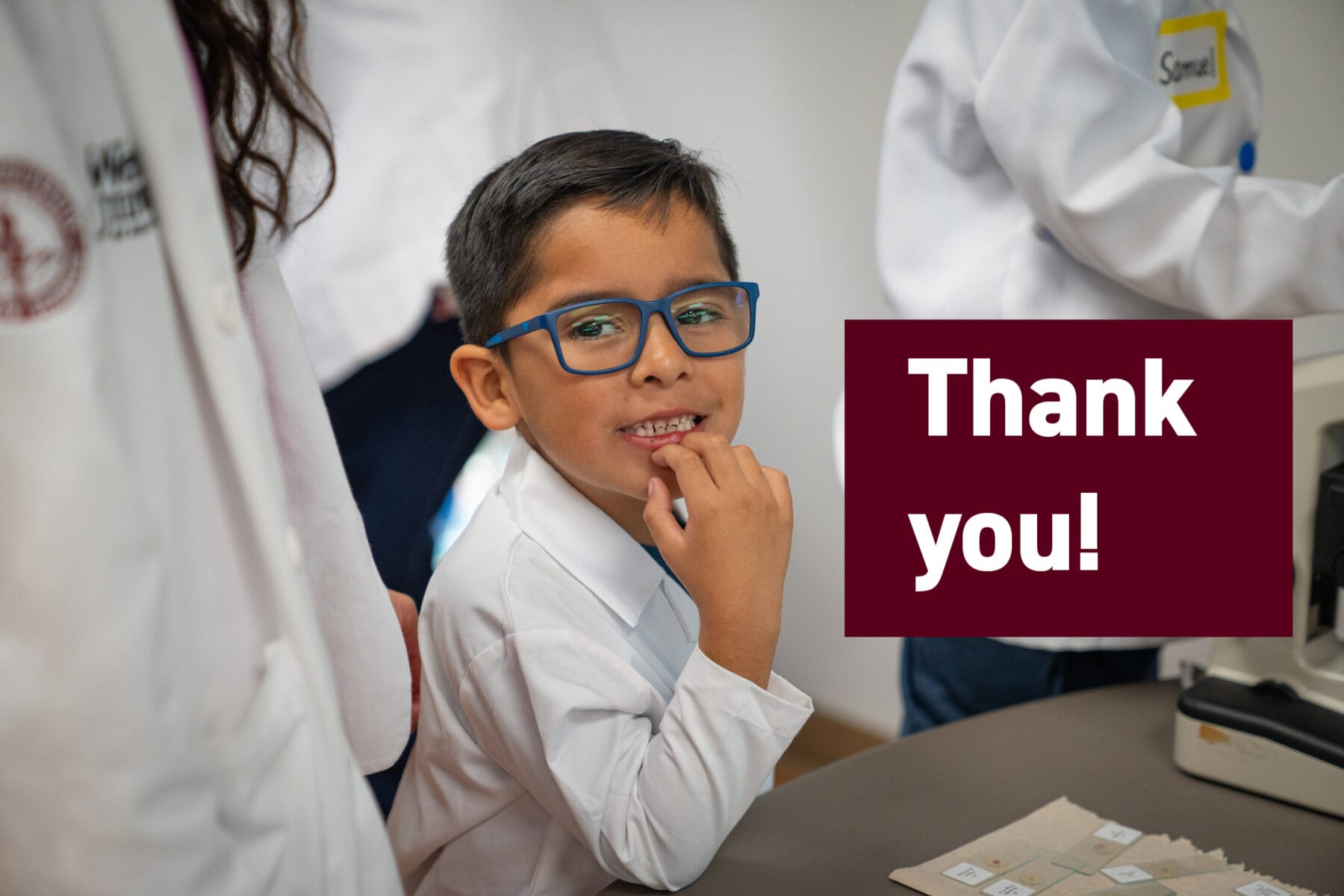 A child wearing glasses and a lab coat smiles while standing next to a table with a microscope. A "Thank you!" sign is in the foreground.
