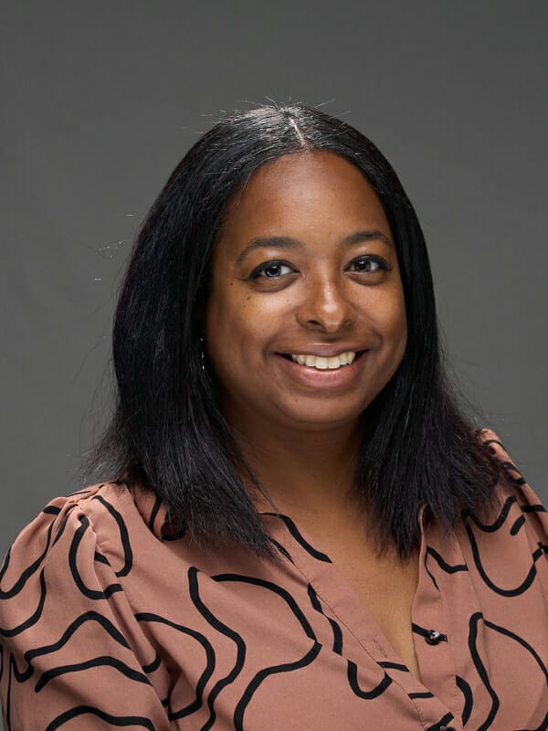 A woman with straight black hair and a patterned brown blouse is smiling at the camera against a plain gray background.