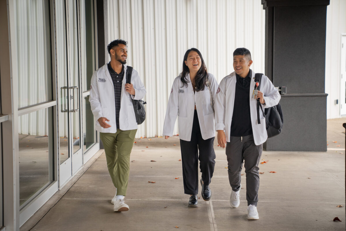 Three students in white lab coats walking down a hallway.