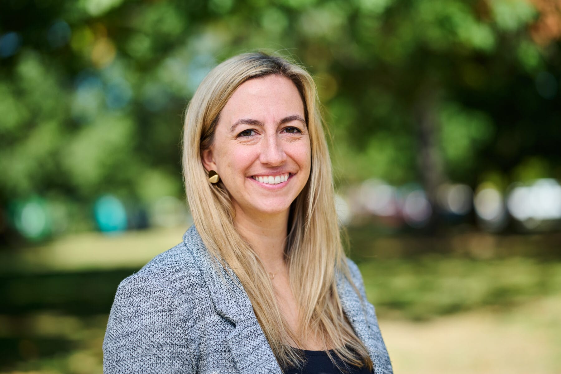 A woman with long blond hair and a gray blazer smiles outdoors with blurred green trees in the background.