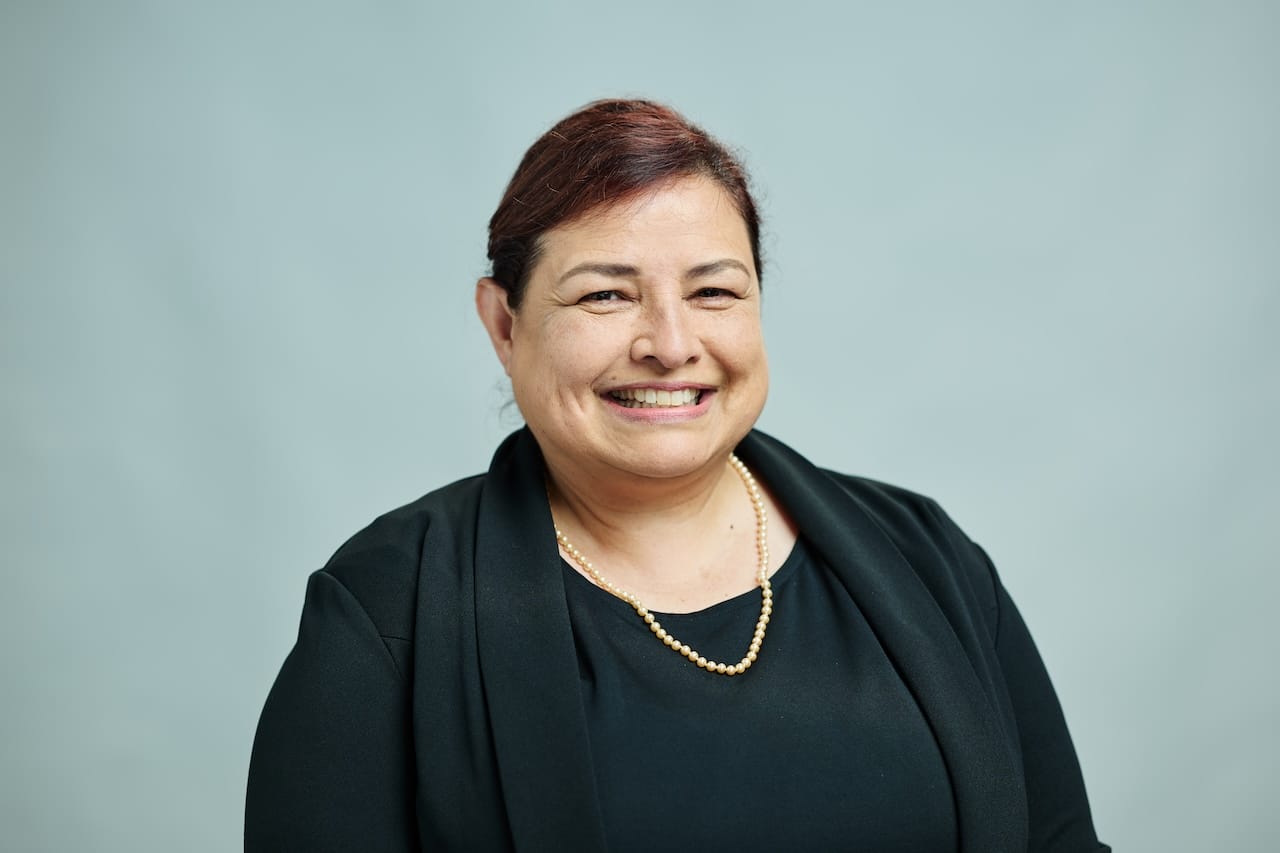 Woman with dark hair and a smile wearing a black top and blazer, with a pearl necklace, in front of a plain background.