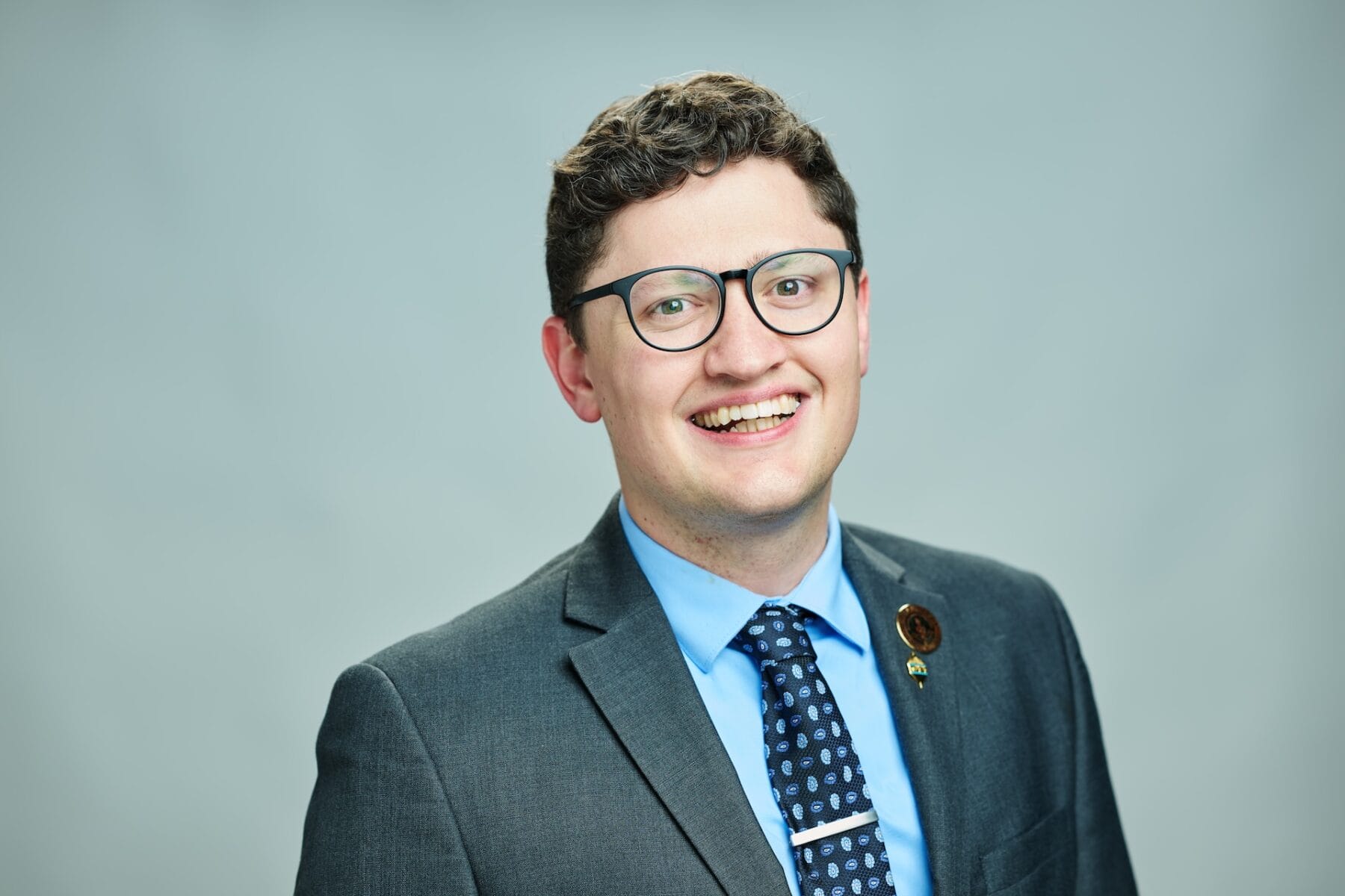 A person wearing a gray suit, blue shirt, patterned tie, glasses, and a lapel pin stands against a plain background, smiling.