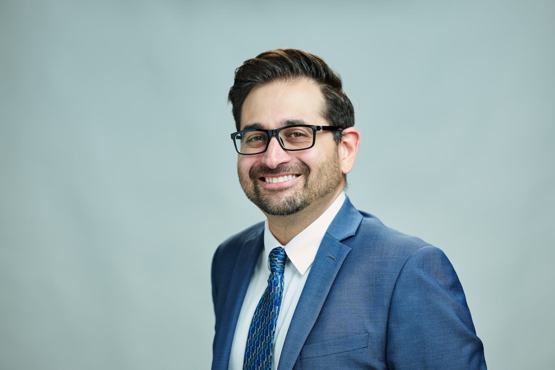 A man wearing glasses and a suit with a blue tie smiles against a plain gray backdrop.