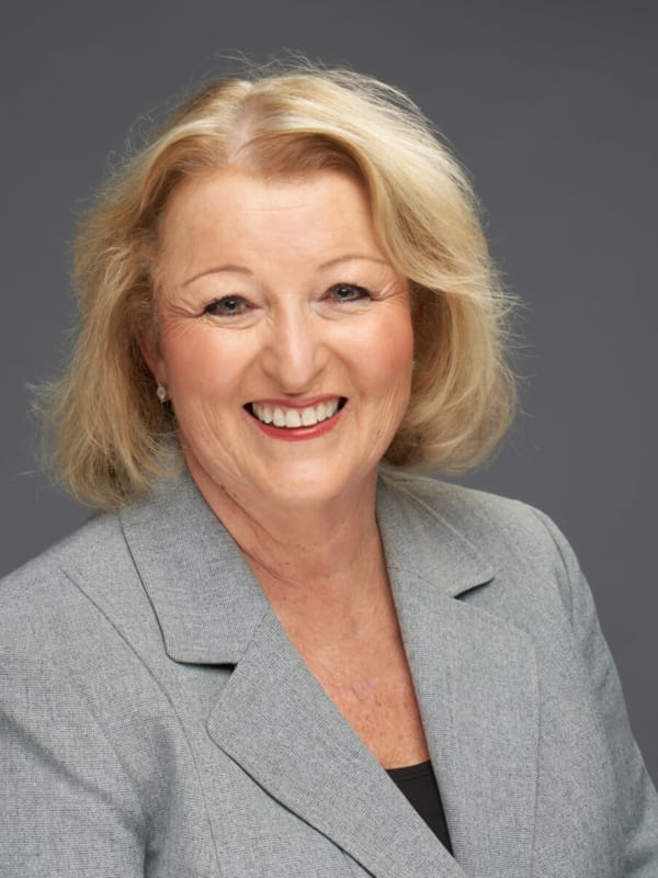 A smiling woman with shoulder-length blonde hair is wearing a gray blazer against a plain gray background.
