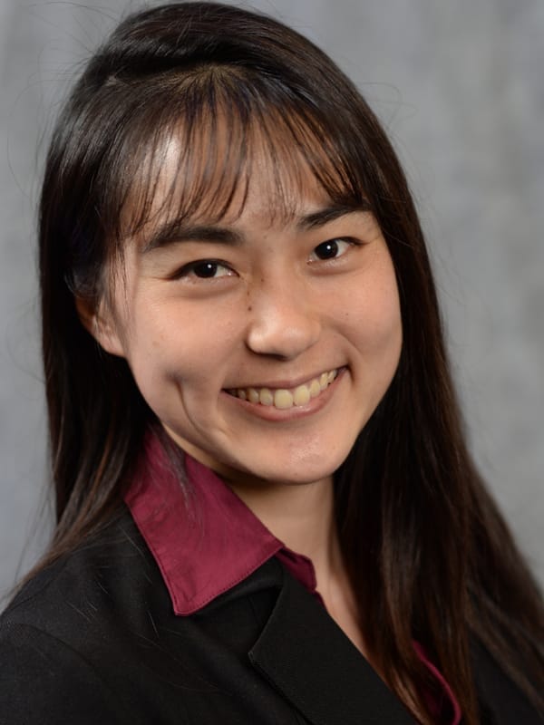A woman with long dark hair and bangs, wearing a dark blazer over a burgundy shirt, smiling at the camera against a neutral gray background.