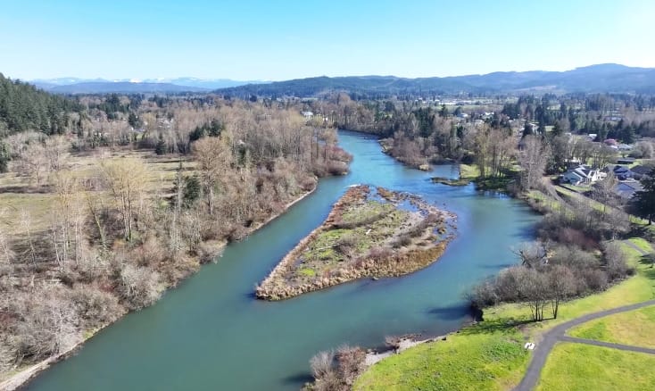 Drone view of a river with an island in the middle, surrounded by green fields and sparse trees, with a town and hills in the background under a clear blue sky.