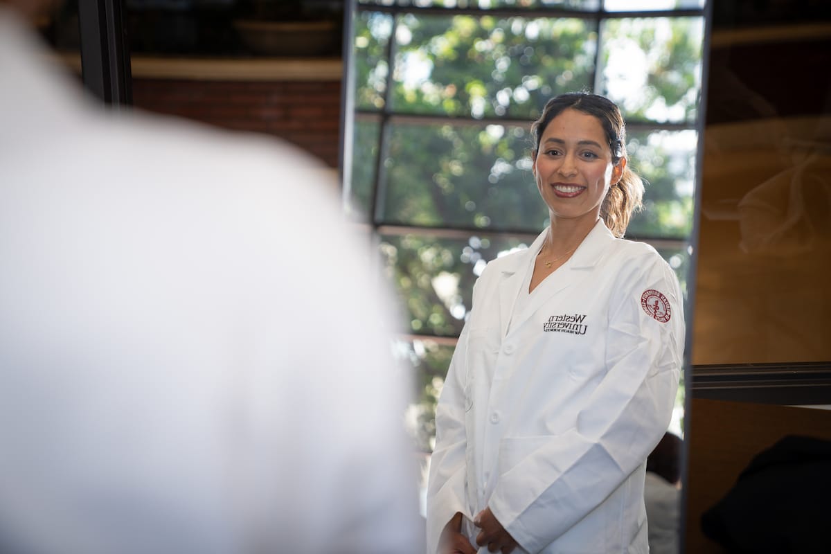 A person in a white lab coat, smiling and looking into a mirror, with trees visible through a window in the background.