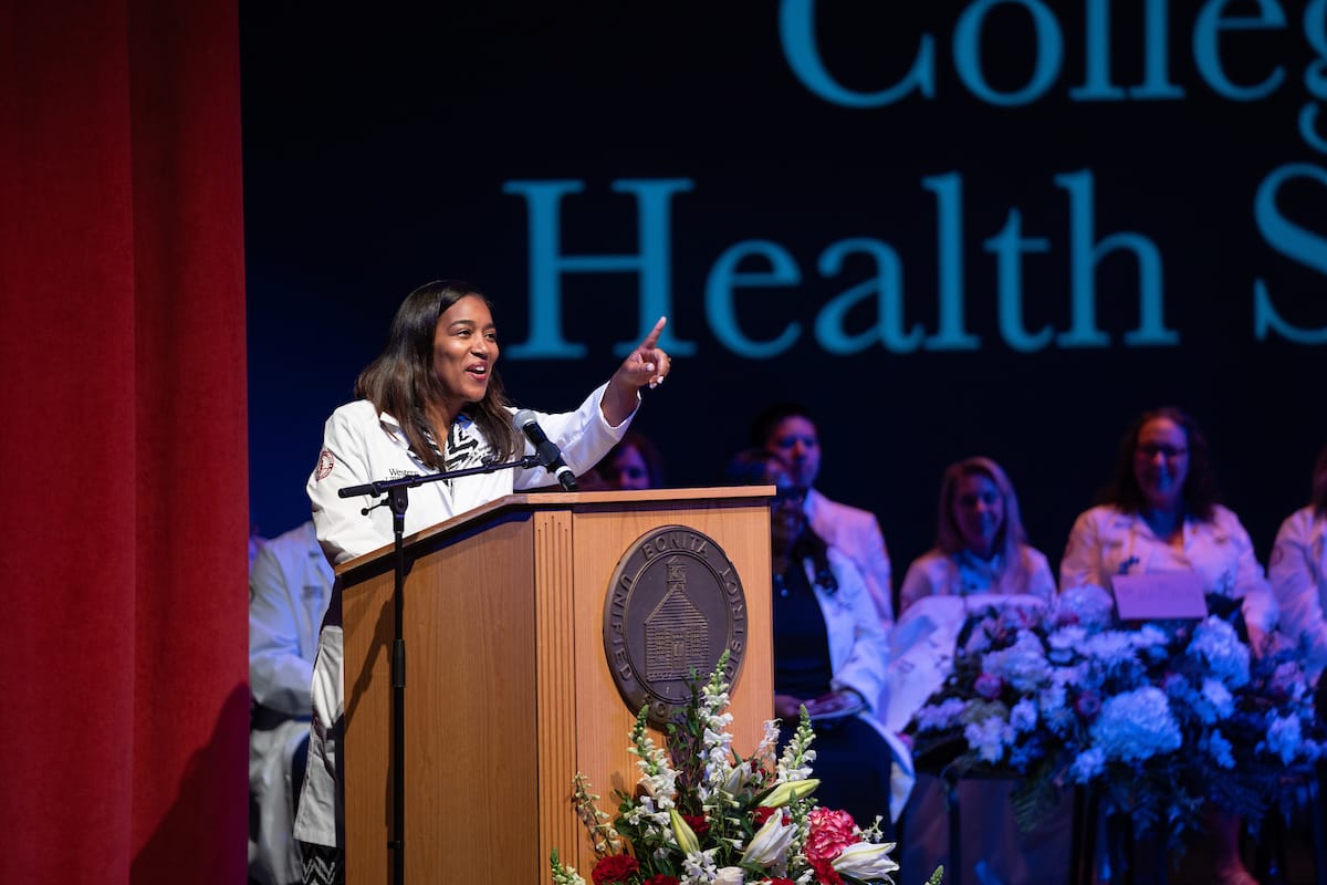 A person in a white coat speaks at a podium with a seal, surrounded by floral arrangements, while others in similar attire sit in the background against a backdrop that reads "College Health S...".