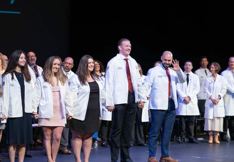 A group of individuals stands on stage wearing white coats, holding hands and smiling, during a ceremony with an audience and other white-coated individuals in the background.