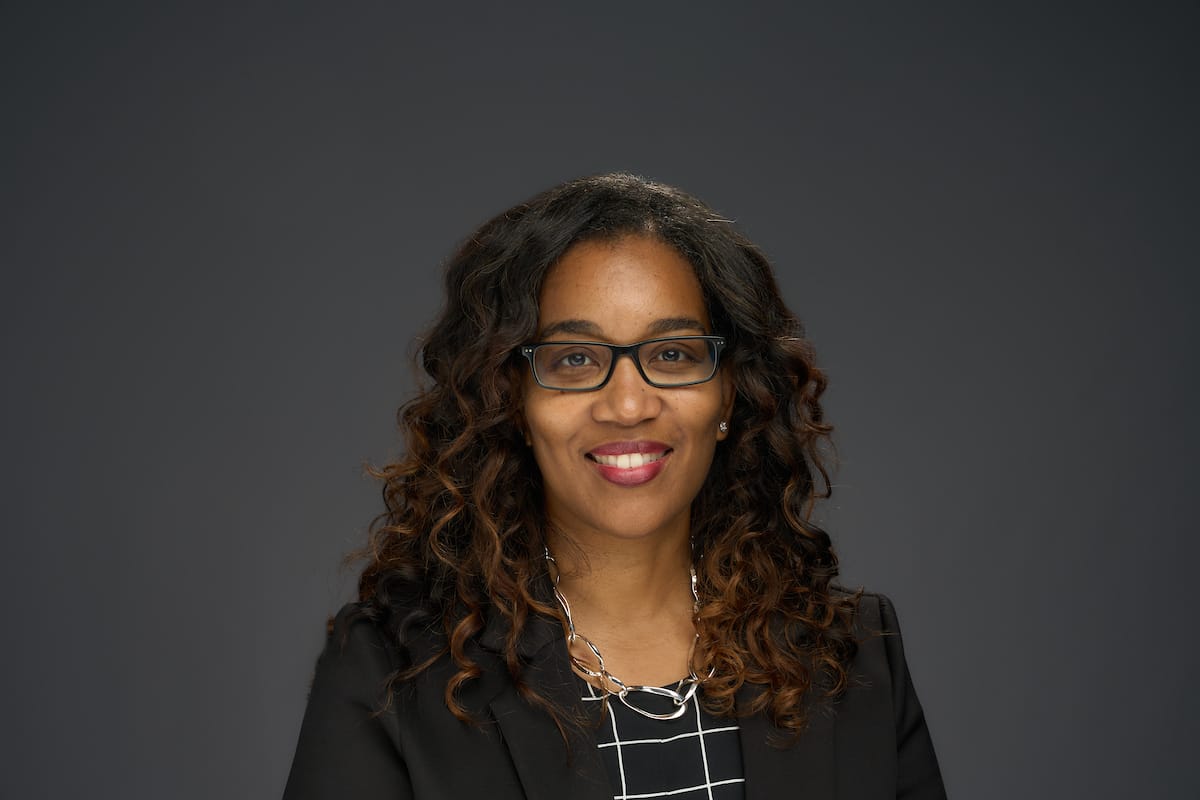 A person with curly hair, wearing glasses, a black blazer, and a patterned top smiles in front of a dark gray background.