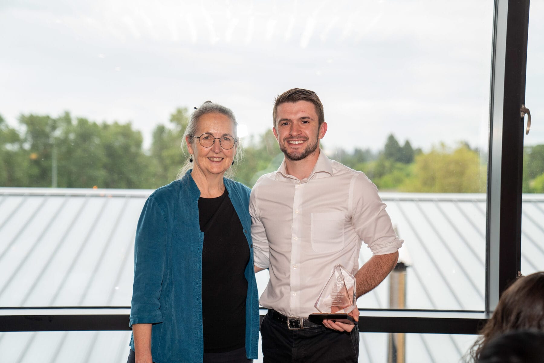 Two people stand indoors by a large window; the person on the left wears glasses and a blue shirt, while the person on the right holds a clear award and smiles.