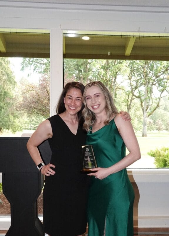 Two women stand indoors, smiling at the camera; one in a black dress, the other in a green dress holding an award. Large windows show trees and greenery outside.