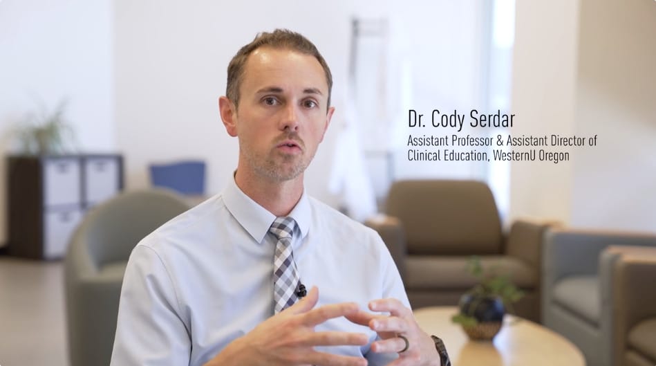A man in a shirt and tie speaks while seated in an office with chairs and shelves in the background. Text on the image identifies him as Dr. Cody Serdar, Assistant Professor and Assistant Director.