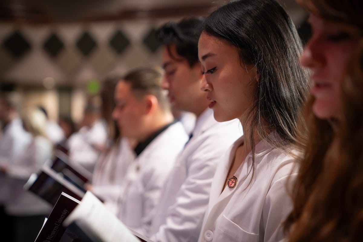 A group of medical students wearing white coats stand in line, attentively reading booklets during a ceremony.