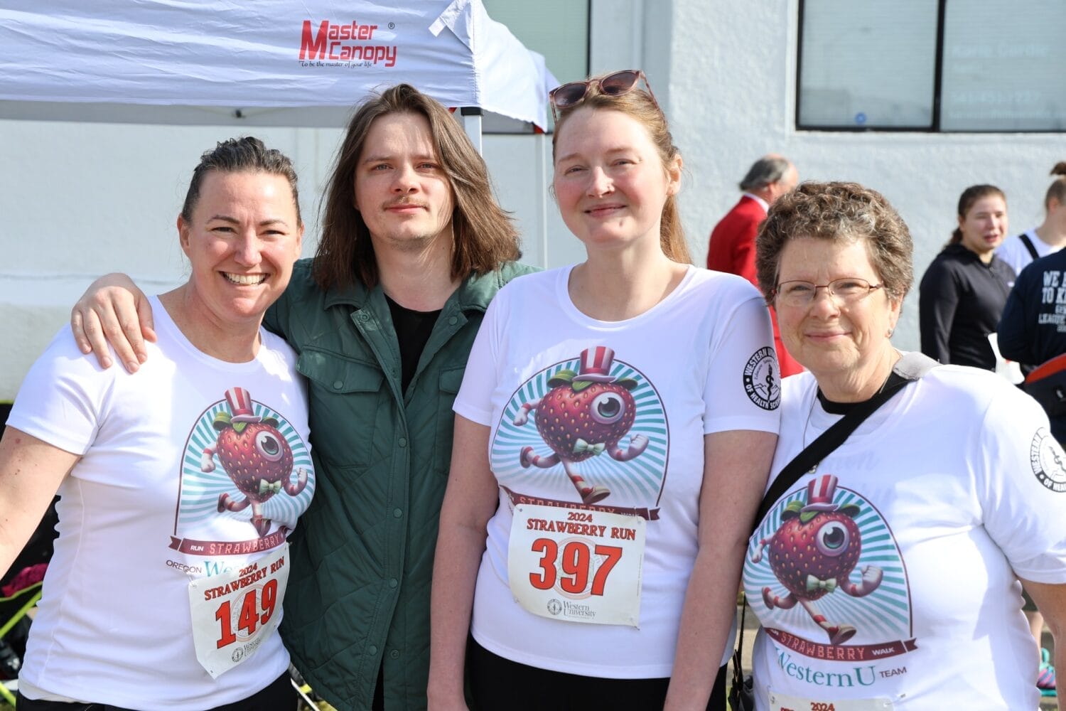 Four people standing together, three wearing matching white shirts with a strawberry design and race numbers, likely participating in a run event. A canopy and other participants are seen in the background.