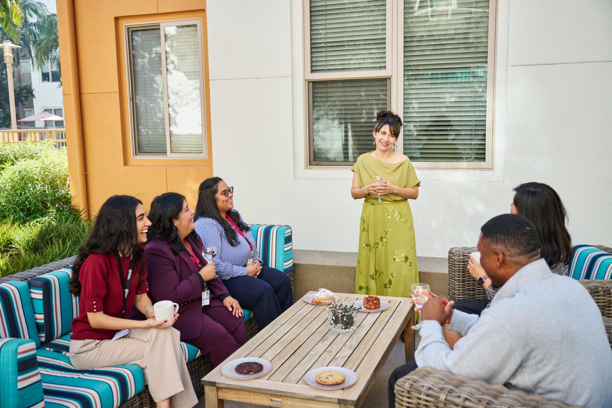 Six people sit and stand around an outdoor coffee table with drinks and snacks, conversing and smiling in a sunlit patio setting.