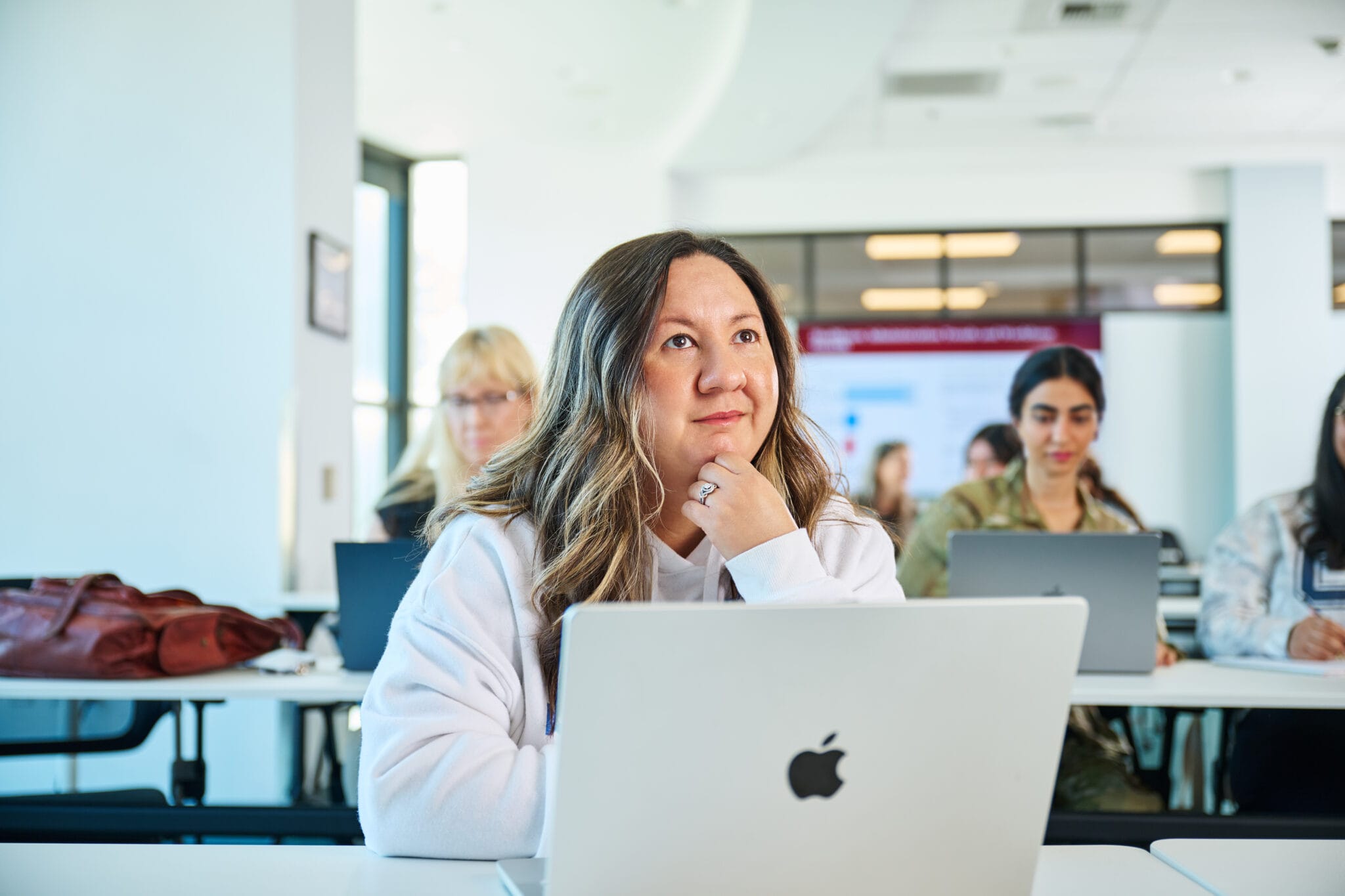 A woman sits at a desk with an Apple laptop in a classroom, looking slightly upward. Other students with laptops are seated in the background.