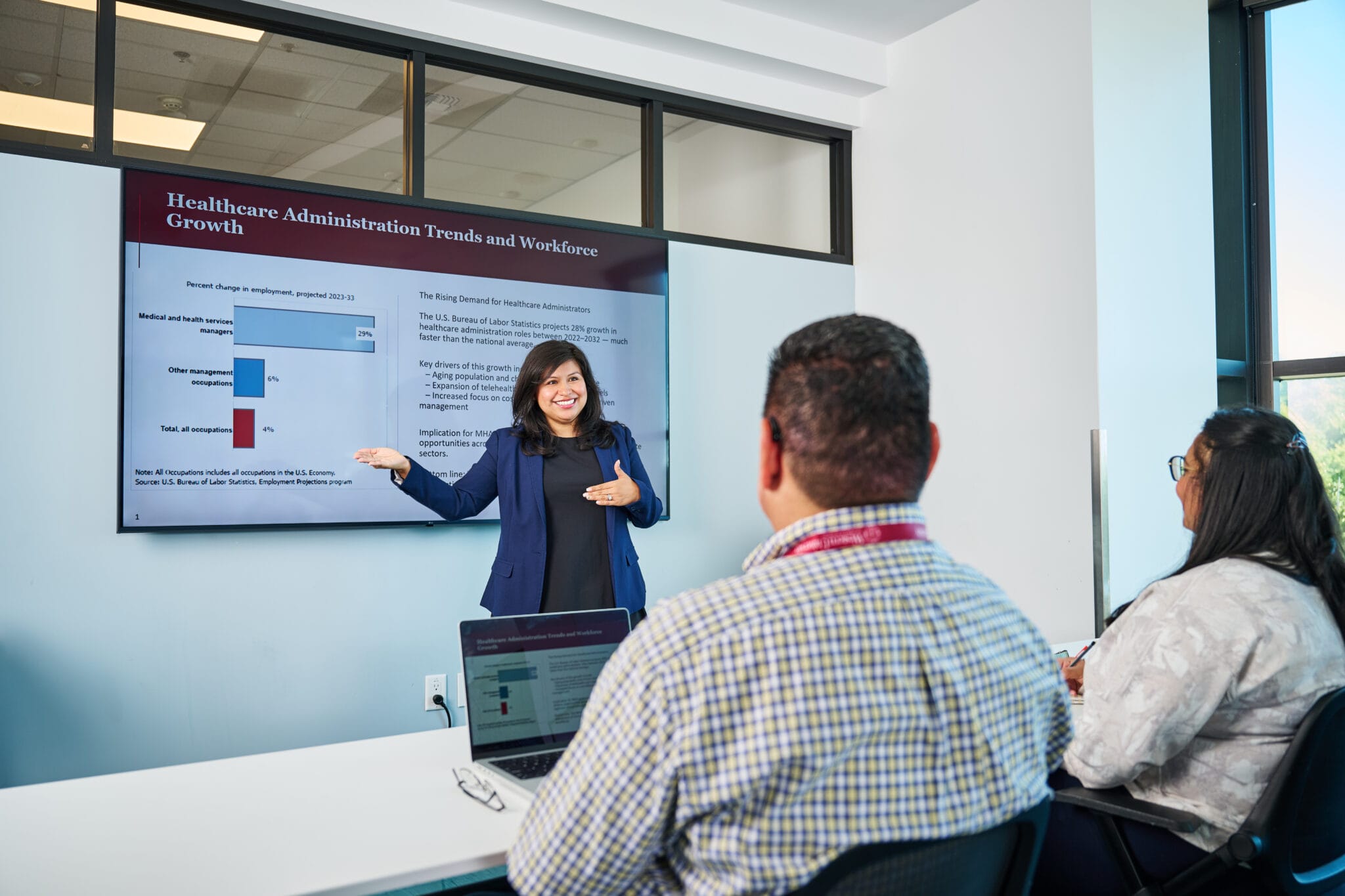 A woman gives a presentation on healthcare administration trends to two seated people in a modern conference room with a large display screen.