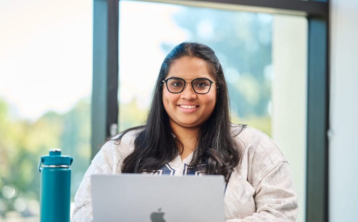 Woman with glasses sits at a desk, smiling at the camera with a laptop open in front of her and a teal water bottle to the side. Large window in the background.