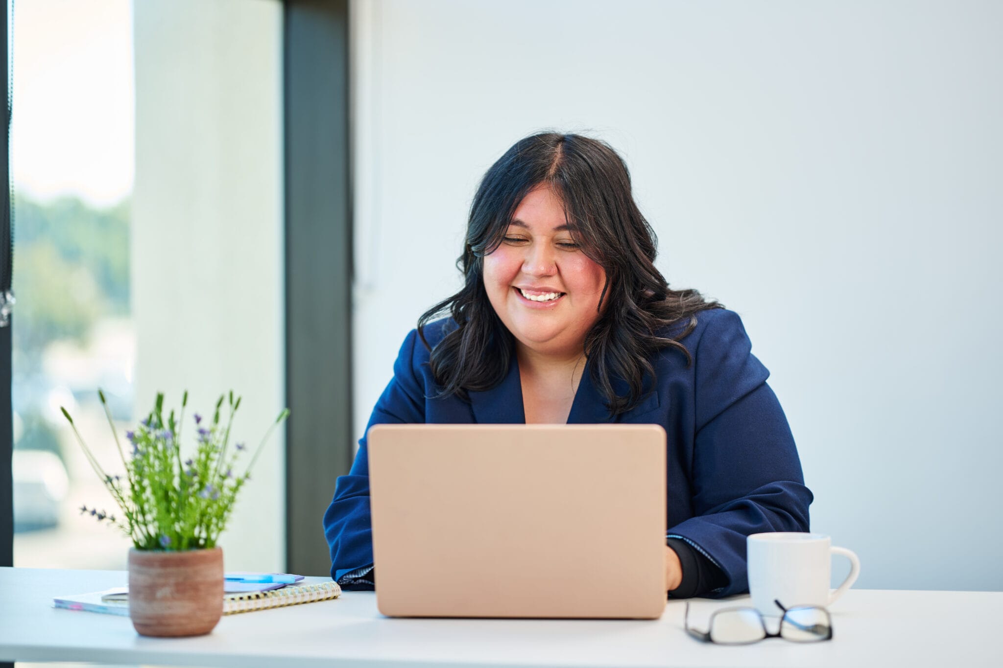 Woman in a blue blazer sits at a desk, smiling at a laptop. A potted plant, notebook, glasses, and a white mug are also on the desk in a bright office.