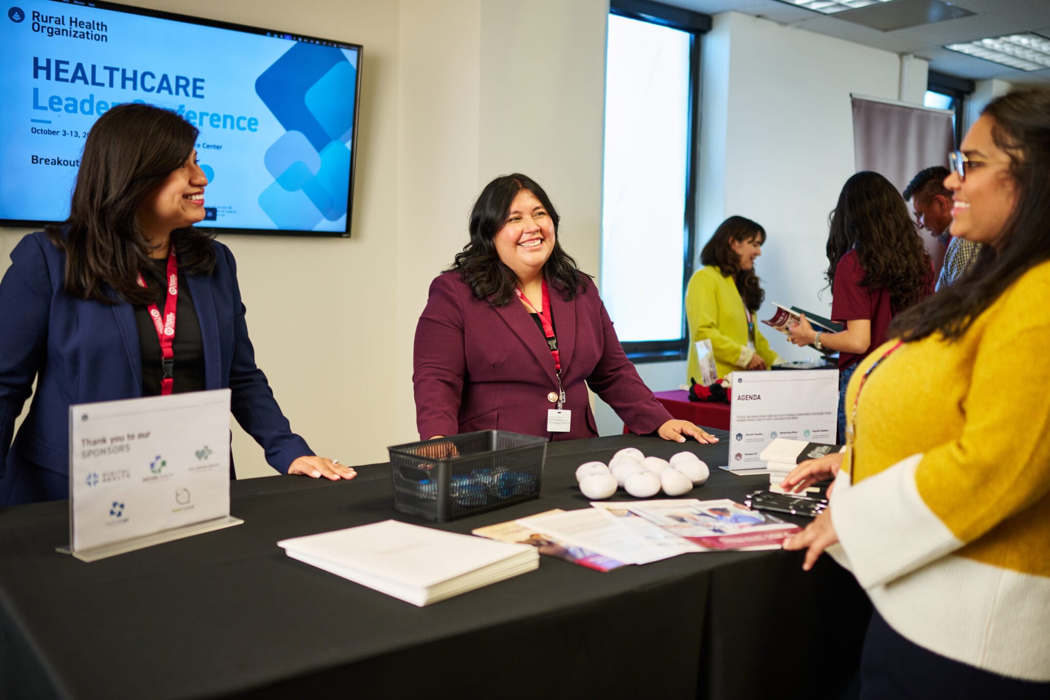 Three women stand and talk at a conference booth with informational materials. A screen in the background displays "Healthcare Leadership Conference.