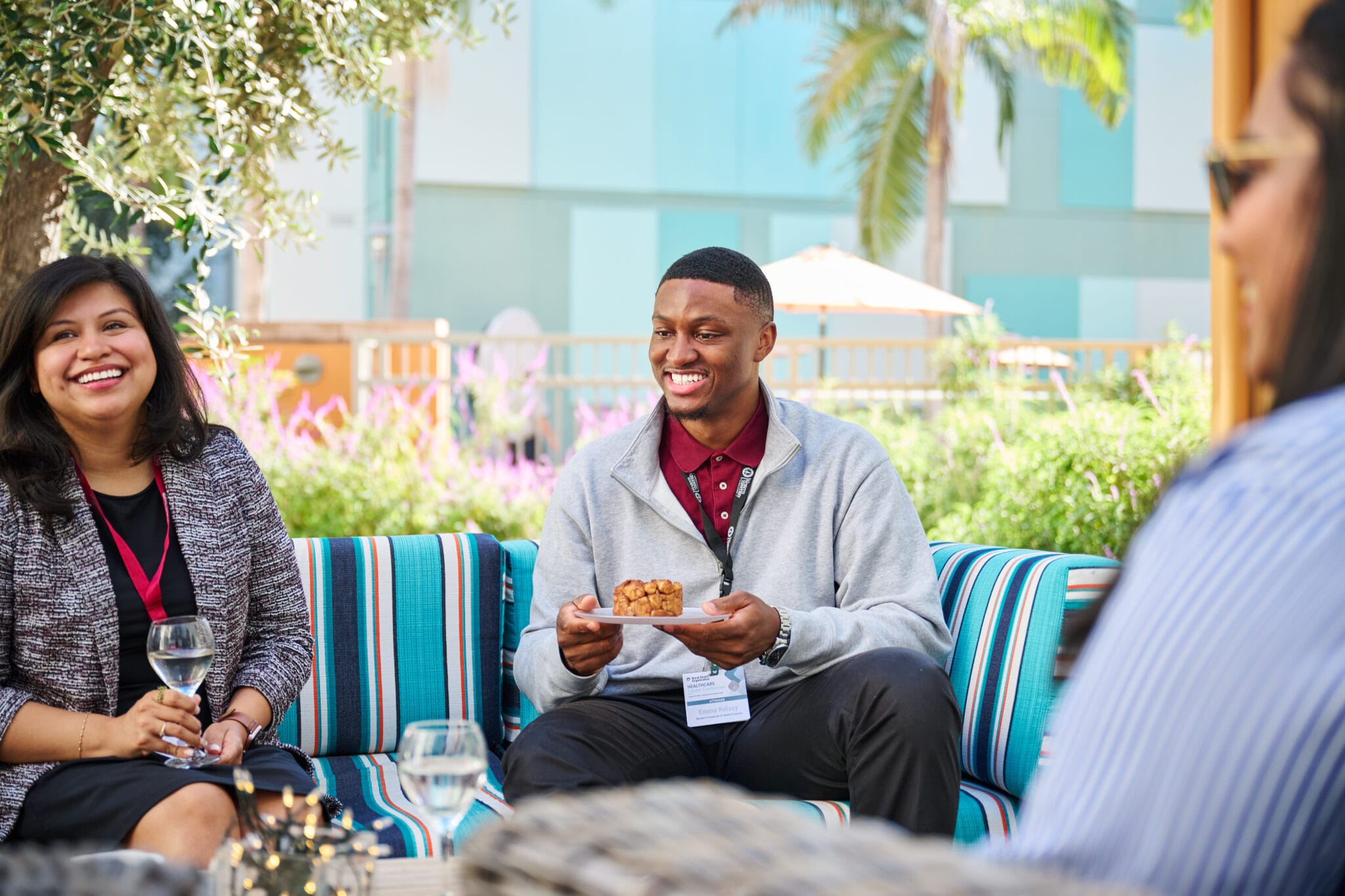 Three people sit on striped outdoor seating, smiling and talking; one person is holding a pastry, others have glasses of water, with greenery and buildings in the background.
