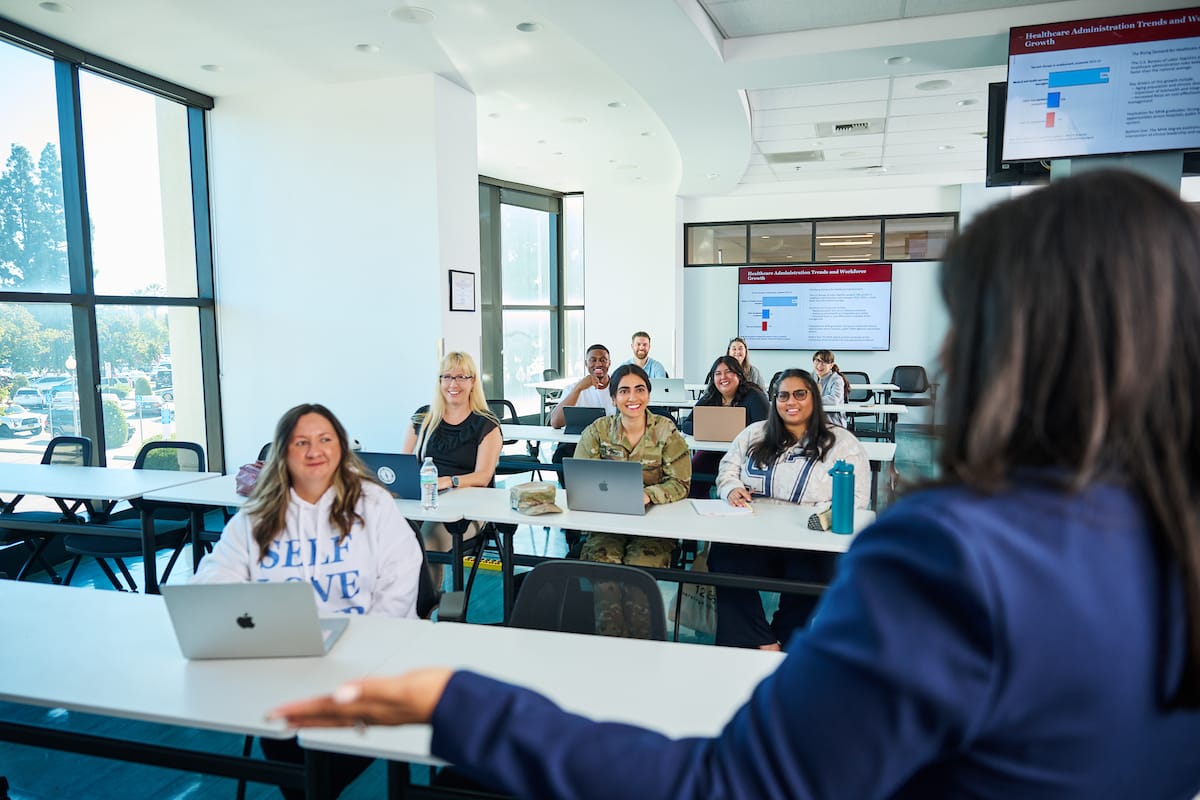 An instructor addresses a diverse group of students seated at desks with laptops in a bright classroom, with presentation slides displayed on screens.