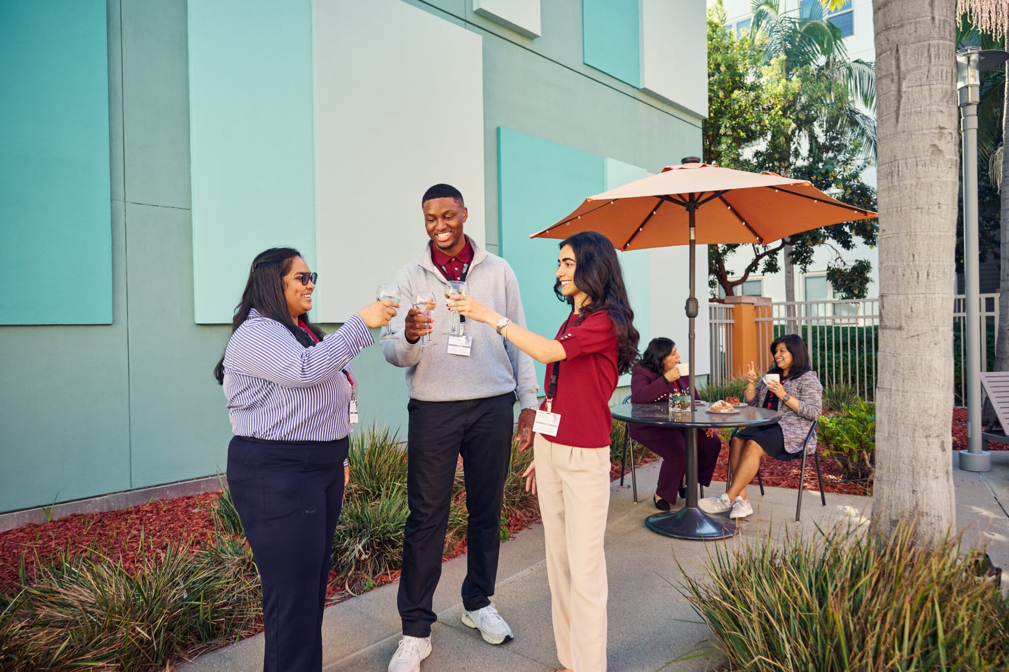 Three people stand outside toasting with drinks, while two others sit and talk at a table under an umbrella in a landscaped courtyard.