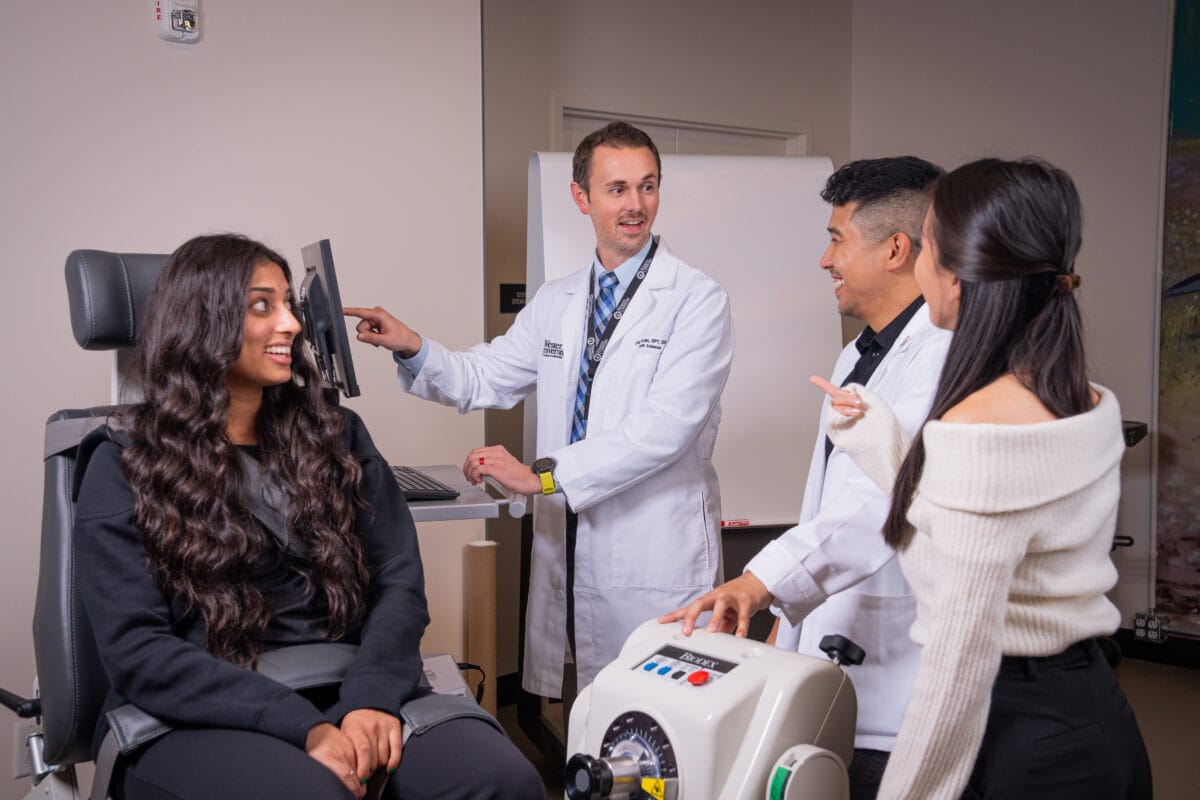 A woman sits in an exam chair while two doctors and another woman stand nearby, discussing something in a medical office.