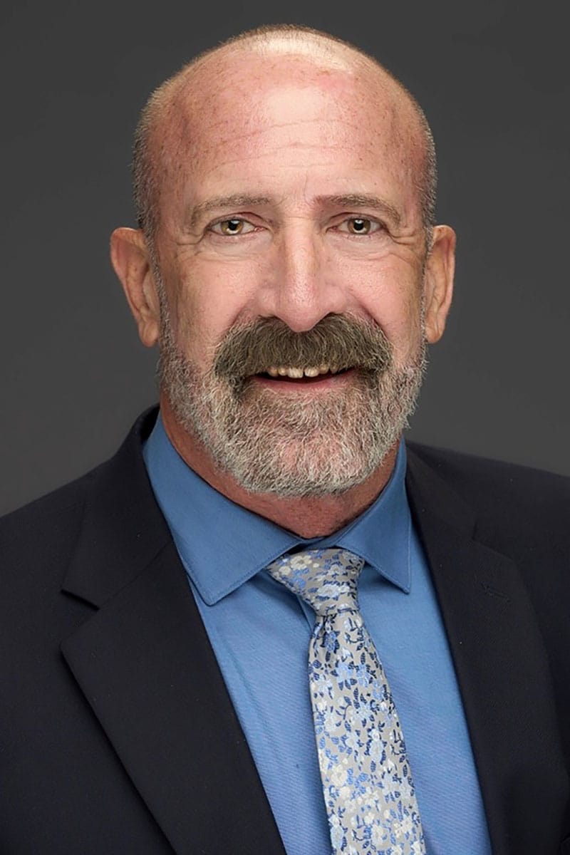 Bald man with a gray beard and mustache wearing a navy suit, blue shirt, and patterned tie, posing against a dark background.